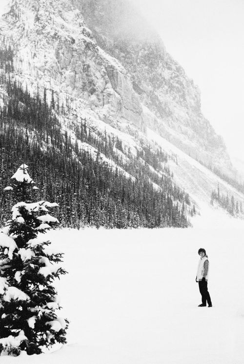 Woman standing on a snow-covered lake with pine tree in the foreground and mountain in the background.