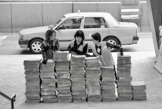 Three ladies standing behind stacks of newspapers to be distributed with a taxi in the background.
