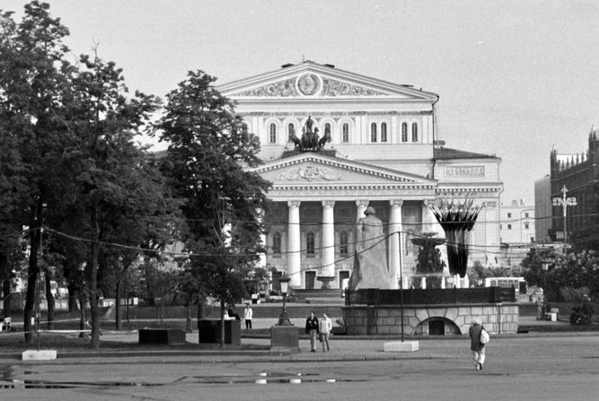 A Greek style building covered by trees on the left third with the back of a Marx statue in foreground.