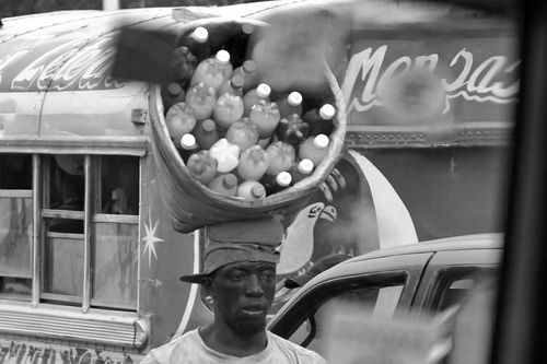 A man carrying a basket of bottles on his head through a busy marketplace.